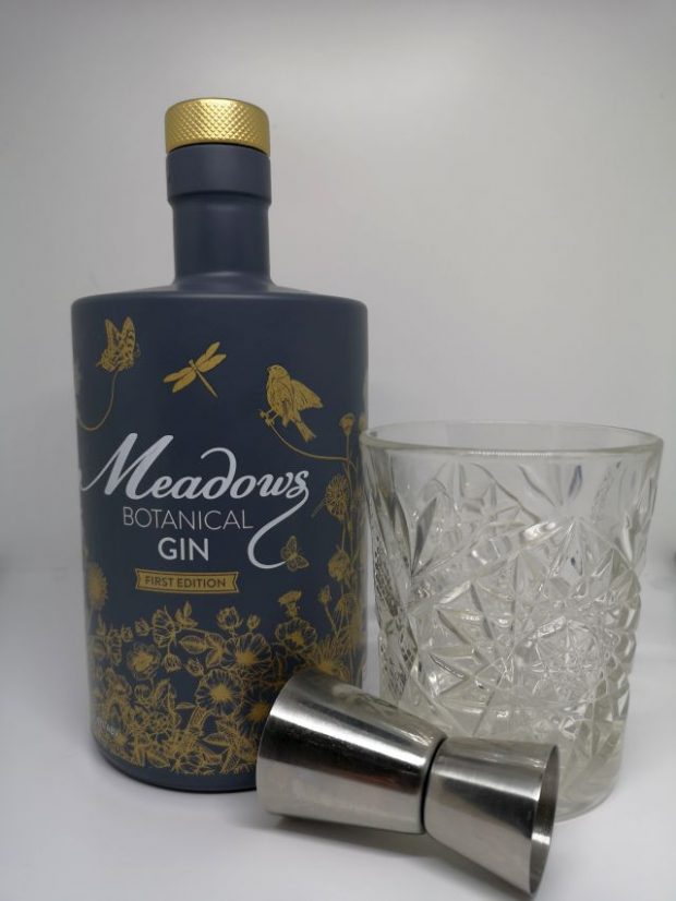 Image of a grey bottle decorated with gold flowers and birds, alongside a silver jigger and a short cut crystal glass containing gin, with a white background.