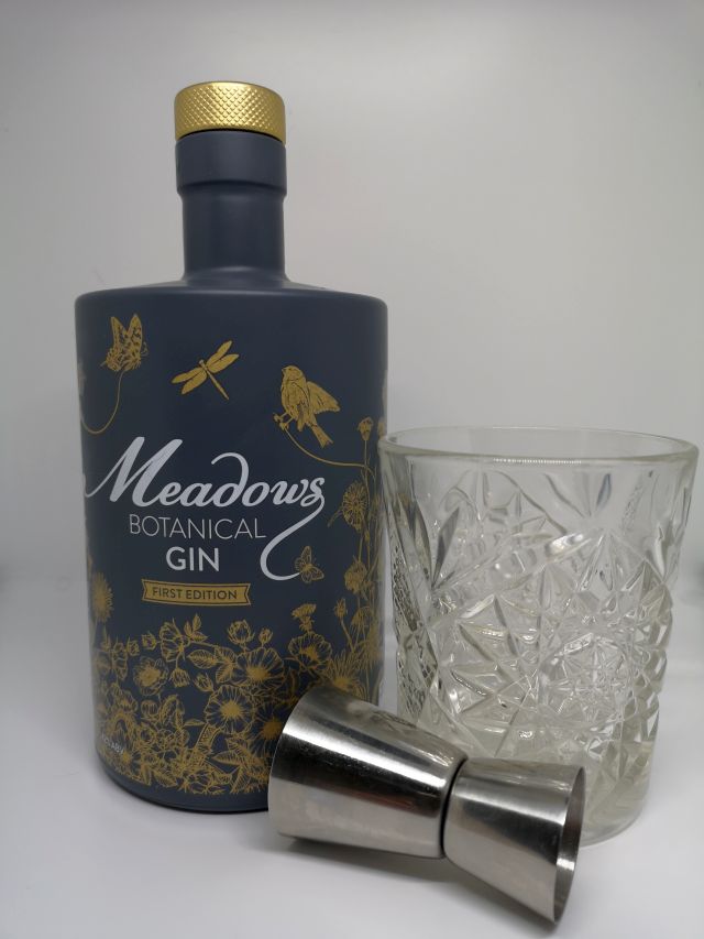 Image of a grey bottle decorated with gold flowers and birds, alongside a silver jigger and a short cut crystal glass containing gin, with a white background.