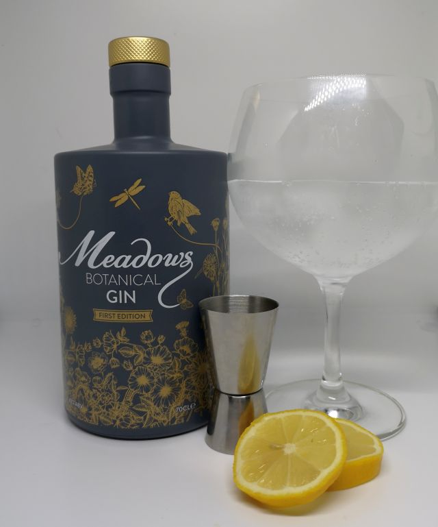Image of a grey bottle decorated with gold flowers and birds, alongside a silver jigger, slices of lemon and a gin copa glass filled with gin tonic and ice, with a white background.