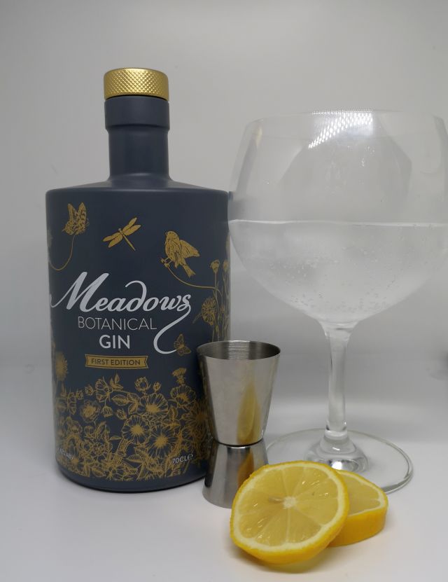 Image of a grey bottle decorated with gold flowers and birds, alongside a silver jigger, slices of lemon and a gin copa glass filled with gin tonic and ice, with a white background.