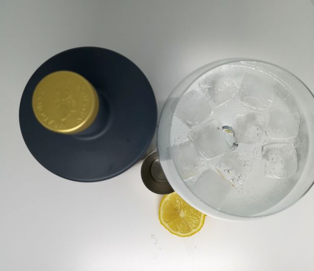 Top down image of a grey bottle with a gold embossed top, next to a silver jigger, slices of lemon and a gin copa glass filled with gin tonic and ice, with a white background.