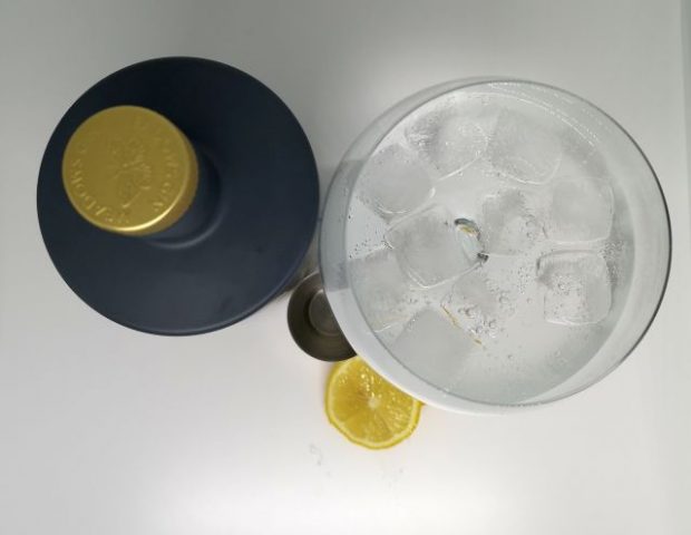 Top down image of a grey bottle with a gold embossed top, next to a silver jigger, slices of lemon and a gin copa glass filled with gin tonic and ice, with a white background.