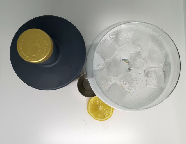 Top down image of a grey bottle with a gold embossed top, next to a silver jigger, slices of lemon and a gin copa glass filled with gin tonic and ice, with a white background.