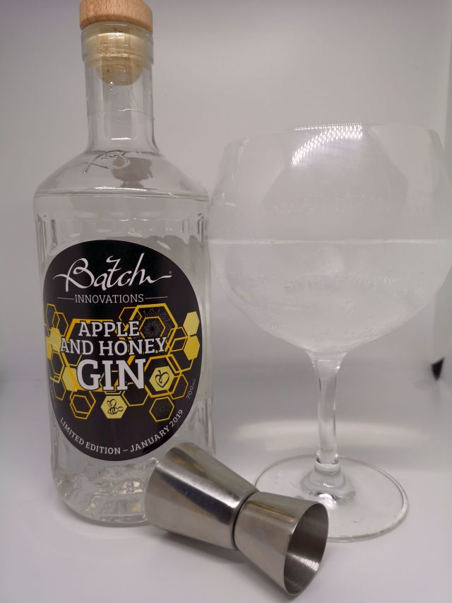 Clear glass bottle of Batch Gin, the label features a honeycome and bees, sitting next to a copa gin glass filled with gin, tonic and ice and a silver jigger
