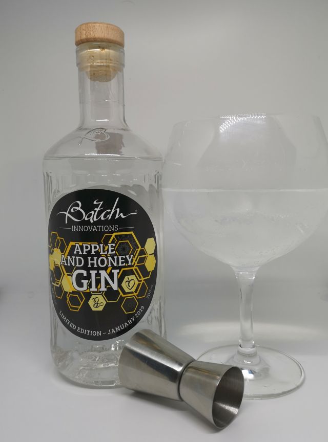 Clear glass bottle of Batch Gin, the label features a honeycome and bees, sitting next to a copa gin glass filled with gin, tonic and ice and a silver jigger