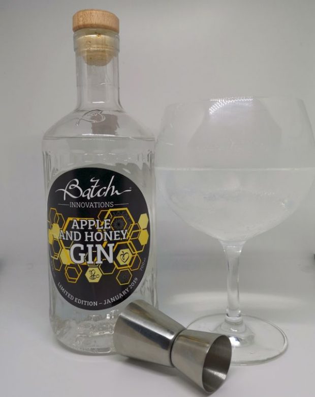Clear glass bottle of Batch Gin, the label features a honeycome and bees, sitting next to a copa gin glass filled with gin, tonic and ice and a silver jigger