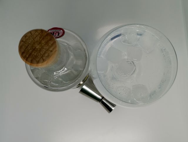 Taken from above, a clear glass bottle of Batch Gin, the label features a honeycome and bees, sitting next to a copa gin glass filled with gin, tonic and ice and a silver jigger