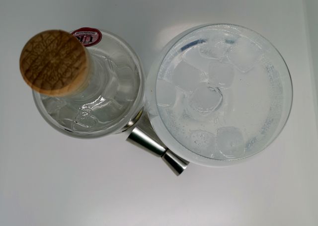 Taken from above, a clear glass bottle of Batch Gin, the label features a honeycome and bees, sitting next to a copa gin glass filled with gin, tonic and ice and a silver jigger