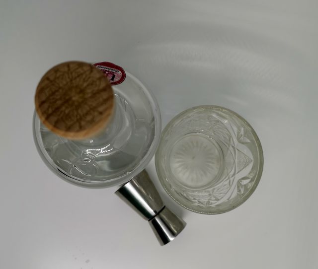Taken from above, a clear glass bottle of Batch Gin, the label features a honeycome and bees, sitting next to a patterned cut glass filled with gin and a silver jigger