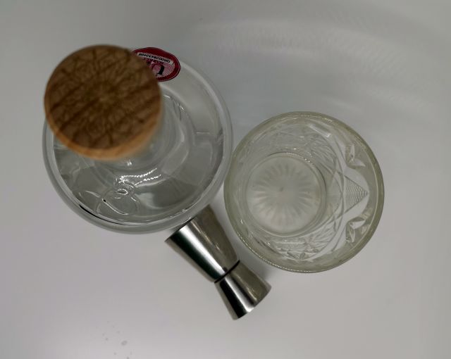 Taken from above, a clear glass bottle of Batch Gin, the label features a honeycome and bees, sitting next to a patterned cut glass filled with gin and a silver jigger